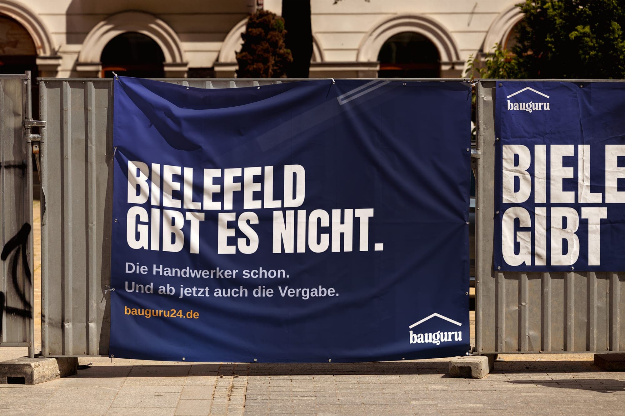 Dunkelblaues BauGuru24-Banner an einem Bauzaun mit der Aufschrift „Bielefeld gibt es nicht. Die Handwerker schon. Und ab jetzt auch die Vergabe.“